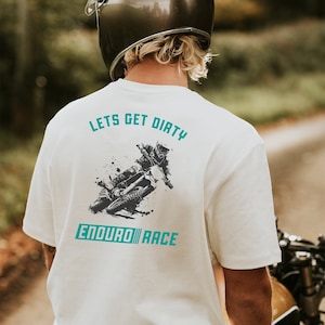 May include: Cream t-shirt featuring a teal graphic of a motocross rider and the words "LET'S GET DIRTY" and "ENDURO RACE". The shirt is worn by a person on a motorcycle, wearing a helmet.