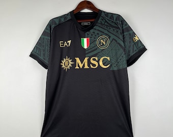 Pode incluir: Camisola de futebol preta com detalhes em verde e dourado. A camisola apresenta um padrão tribal, as letras "MSC", um sol com um círculo no seu interior e a bandeira italiana. O texto "EA7" está no lado esquerdo da camisola e a letra "N" no lado direito.