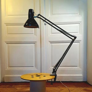 Puede incluir: Una lámpara de escritorio negra articulada con una pantalla cónica y un brazo largo y ajustable. La lámpara está sujeta a una mesa redonda de madera clara. La mesa se apoya sobre una base cilíndrica. El diseño de la lámpara es industrial.