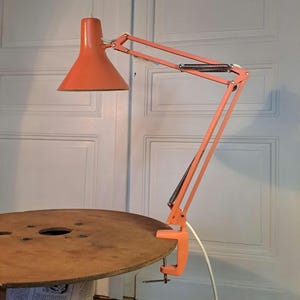 Puede incluir: Lámpara de escritorio naranja con brazo ajustable y base de abrazadera. La lámpara tiene una pantalla cónica y un cable de alimentación blanco. La lámpara está sujeta a una bobina de madera.