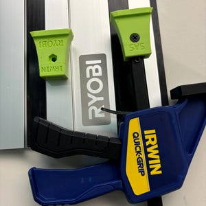 Könnte beinhalten: Eine blaue und gelbe Irwin Quick-Grip-Zwinge mit einem schwarzen Griff. Die Zwinge ist an einer silbernen Schiene mit zwei grünen Kunststoffaufsätzen befestigt, die mit "IRWIN" und "RYOBI" beschriftet sind.