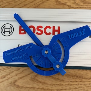 Puede incluir: Una herramienta de transportador azul con el texto "TOOLAX" y marcas de grados. La herramienta está diseñada para ser compatible con los productos Bosch, como indica el texto "Compatible con! BOSCH ®". La herramienta está sobre una superficie blanca y plateada.