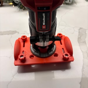 Könnte beinhalten: Ein Einhell-Akku-Fräser in Rot und Grau mit roter Basis und Tiefenanschlag. Der Fräser steht auf einer weißen Oberfläche.