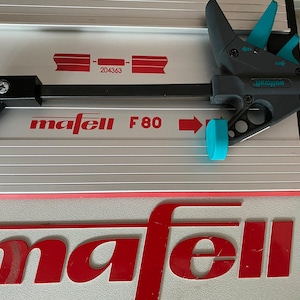 Puede incluir: Primer plano de una sierra de mesa mafell F80 plateada y roja con una abrazadera negra y azul. La abrazadera está etiquetada como "Wolfcraft".