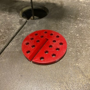 May include: A red plastic jig with multiple holes, designed for use with a scroll saw. The jig is split into two halves, with a slot in the middle.