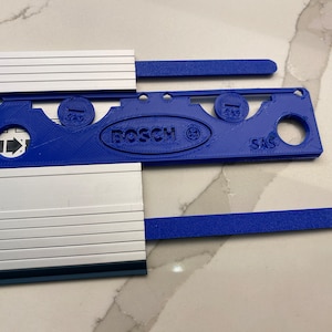 Op de afbeelding: Een blauwe plastic tool met het Bosch-logo en de woorden "SAS" en "GAS" erop gedrukt. De tool wordt gebruikt voor het instellen van een zaag.