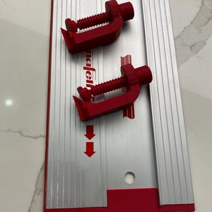 May include: Two red clamps are attached to a silver metal ruler with red markings. The ruler has a hole in the bottom right corner.