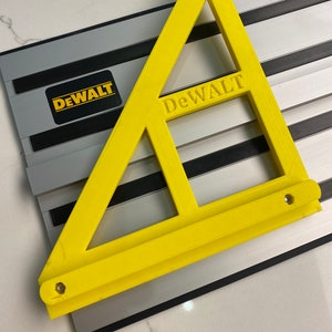 Puede incluir: Una regla triangular de plástico amarillo con la palabra "DEWALT" impresa en ella. La regla está encima de un sistema de rieles plateado y negro.