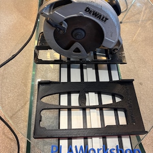 May include: A DeWalt circular saw with a yellow and silver body, resting on a black and white surface. A black plastic guide and blue "PLAWorkshop" text are in the foreground. The saw blade is visible.
