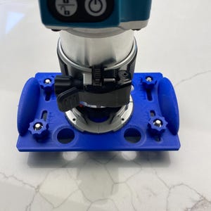 May include: A blue router jig with a router mounted on top. The jig has several holes and knobs for adjusting the router's position.