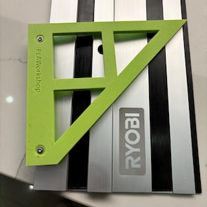 Peut inclure: Une règle triangulaire en plastique vert avec le texte "PLAYworkshop" imprimé dessus. La règle est placée sur un système de rails argenté et noir avec le mot "RYOBI" imprimé dessus.