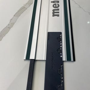 Puede incluir: Un riel guía de metal plateado y verde con una barra guía de plástico negro. El riel guía tiene el texto "metabo" impreso en él. La barra guía tiene el texto "guide rail" impreso en él.