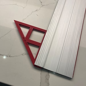 Peut inclure: Une règle triangulaire en plastique rouge de 45 degrés avec le texte "45 Degree" et "Flexi-Ruler" imprimé dessus. La règle est placée sur un comptoir blanc.