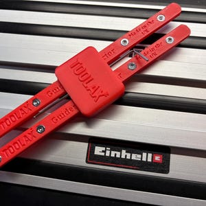 Può includere: Un utensile di guida TOOLAX rosso con la scritta "Made in the UK" e "Guide" stampata sopra. L'utensile è su una superficie a strisce argento e nere. Il marchio "Einhell" è visibile.