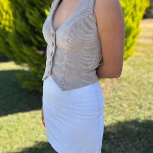 May include: A sleeveless beige corduroy vest featuring a V-neckline, a front button closure, and a pointed hemline, paired with a plain white mini skirt. The vest has a vertical ribbed texture and is fitted to the torso with visible seams.