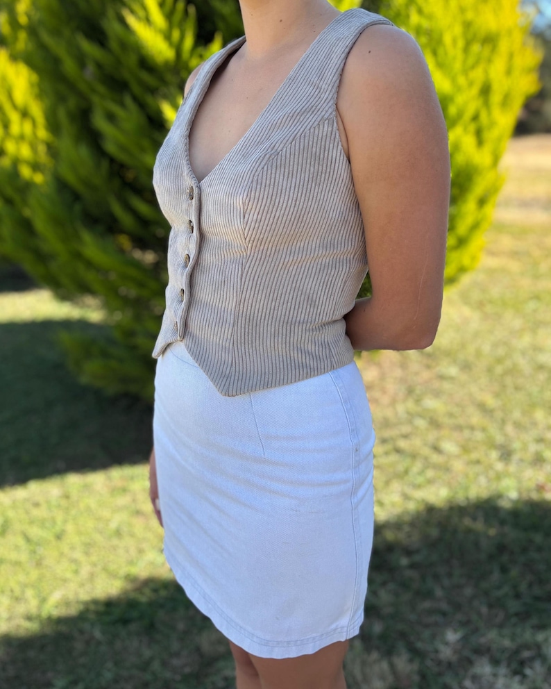 May include: A sleeveless beige corduroy vest featuring a V-neckline, a front button closure, and a pointed hemline, paired with a plain white mini skirt. The vest has a vertical ribbed texture and is fitted to the torso with visible seams.