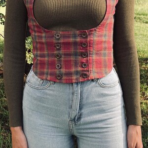 K&ouml;nnte beinhalten: Ein olivgr&uuml;nes, geripptes Langarmshirt mit Rollkragen unter einer rot-blau karierten Korsett-Weste mit zwei vertikalen Reihen brauner Kn&ouml;pfe und breiten Schultertr&auml;gern, kombiniert mit einer hellblauen Jeans mit hoher Taille.