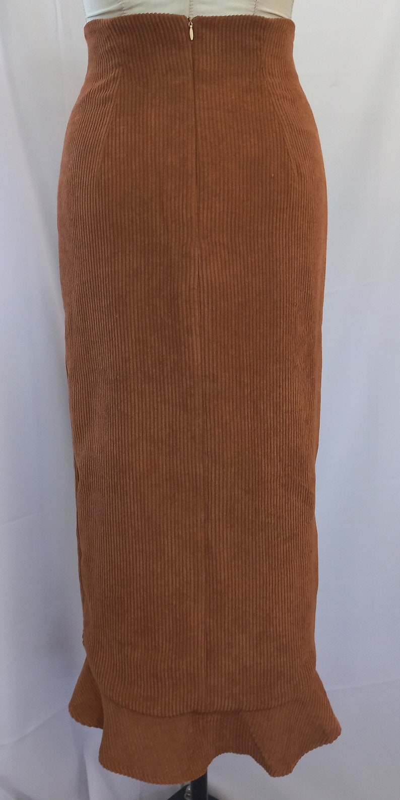 May include: A brown corduroy skirt with a high waist and a slight flare at the hem. The skirt has a vertical ribbed texture and a zipper closure in the back. The skirt is a solid color and appears to be made of a soft fabric.