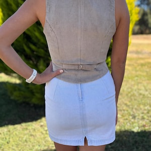May include: A beige corduroy vest featuring a vertical ribbed texture and a rear adjustable strap with a metal buckle, paired with a light blue denim mini skirt that has a central vertical seam and a small slit at the bottom hemline.