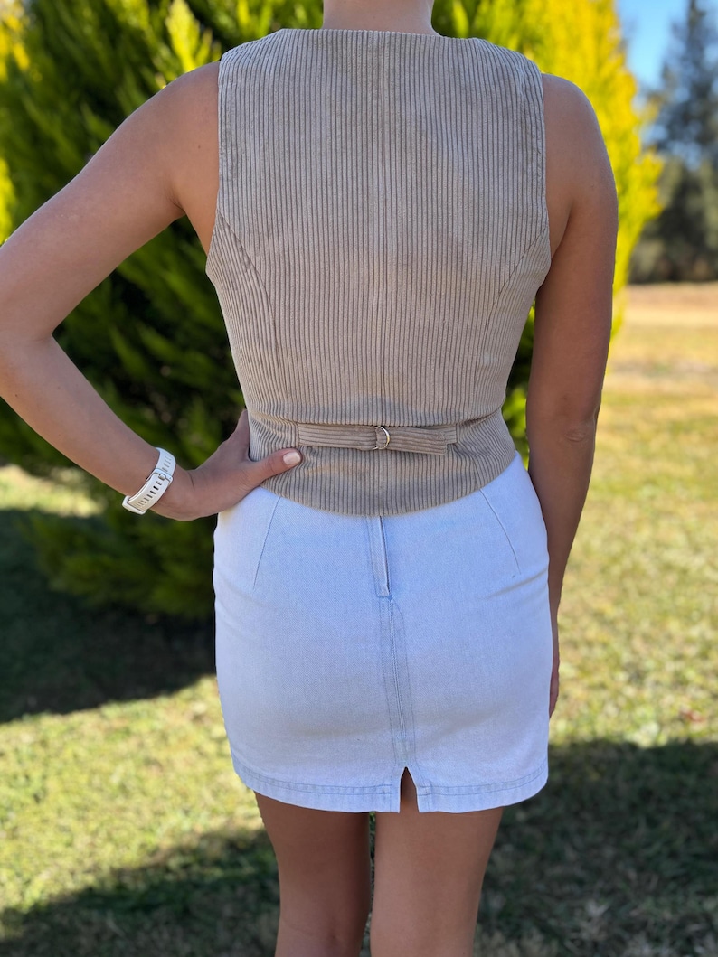 May include: A beige corduroy vest featuring a vertical ribbed texture and a rear adjustable strap with a metal buckle, paired with a light blue denim mini skirt that has a central vertical seam and a small slit at the bottom hemline.