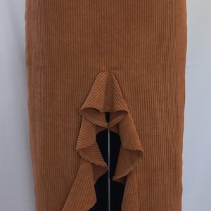 May include: A rust-colored corduroy skirt with a high waist and a ruffled asymmetrical hem. The skirt is displayed on a mannequin and has vertical ribbing. The skirt's design includes a unique ruffle detail.