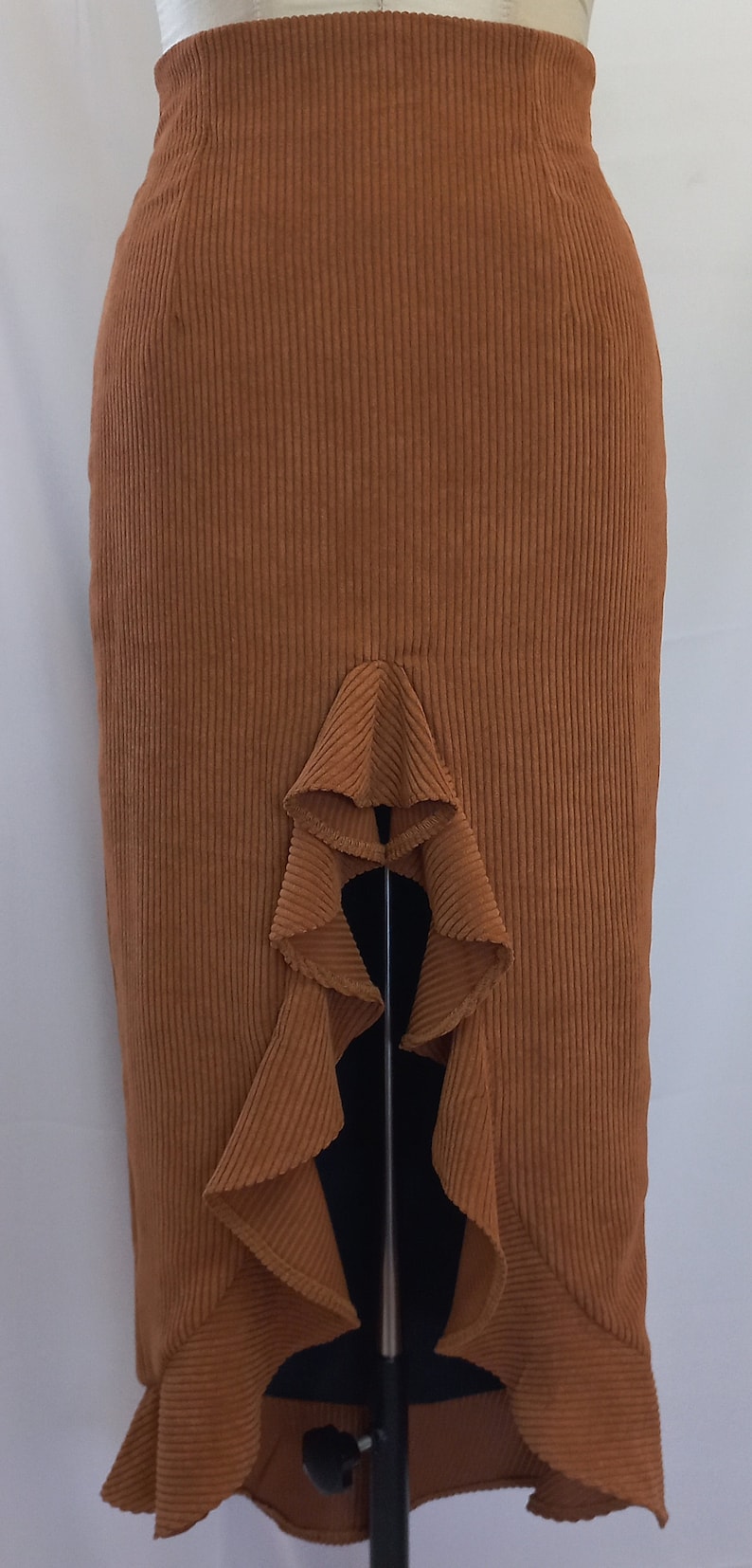 May include: A rust-colored corduroy skirt with a high waist and a ruffled asymmetrical hem. The skirt is displayed on a mannequin and has vertical ribbing. The skirt's design includes a unique ruffle detail.