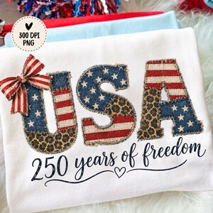 Op de afbeelding: Wit T-shirt met het woord "USA" in een patriottisch ontwerp, met sterren, strepen en een luipaardprint. De tekst "250 years of freedom" staat eronder. Een rood-wit-blauwe strik staat linksboven.