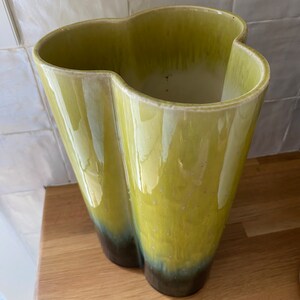 May include: A tall, ceramic vase with a unique, wavy shape. The vase is a pale yellow-green color, with a darker, brown-black gradient at the base. The interior is white. The vase is sitting on a wooden surface.