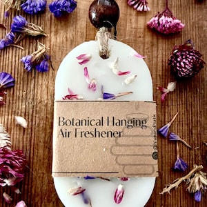 May include: A white, oval-shaped botanical hanging air freshener with the word "Sweetpea" at the bottom. The freshener is adorned with dried flower petals and is suspended by a twine cord and a wooden bead. The background is a wooden surface.