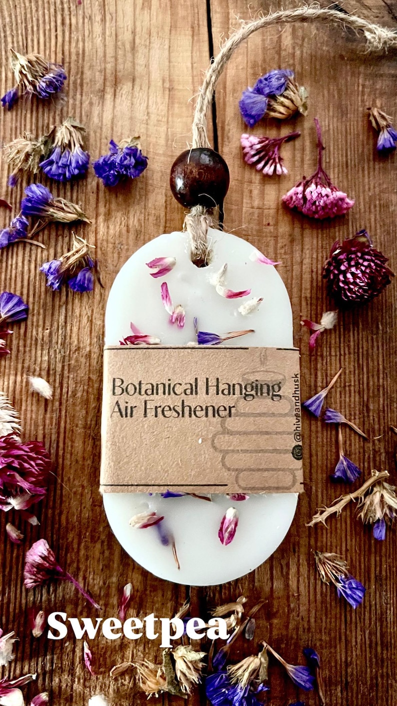 May include: A white, oval-shaped botanical hanging air freshener with the word "Sweetpea" at the bottom. The freshener is adorned with dried flower petals and is suspended by a twine cord and a wooden bead. The background is a wooden surface.