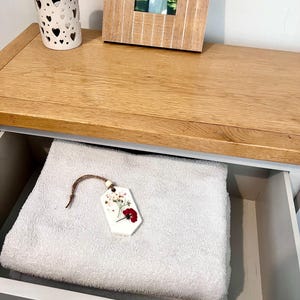 May include: A white ceramic candle holder with heart-shaped cutouts sits on a wooden surface. A framed photo and a drawer with a folded white towel and a floral-decorated tag are also visible. The drawer is painted gray.