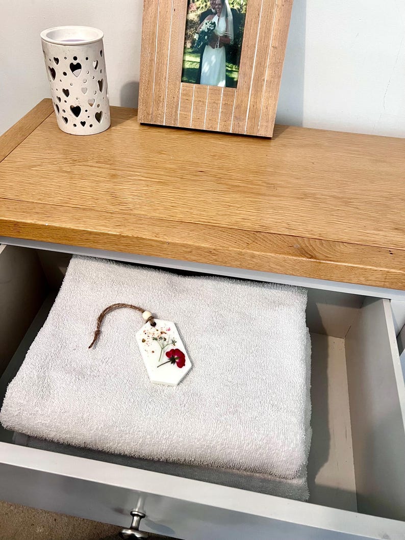 May include: A white ceramic candle holder with heart-shaped cutouts sits on a wooden surface. A framed photo and a drawer with a folded white towel and a floral-decorated tag are also visible. The drawer is painted gray.