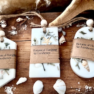 May include: Three white botanical hanging air fresheners with natural elements like shells and dried plants. Each has a brown paper label with the text "Botanical Hanging Air Freshener" and is suspended by a string with a wooden bead.