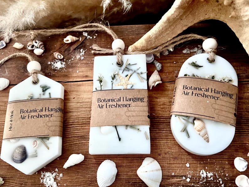 May include: Three white botanical hanging air fresheners with natural elements like shells and dried plants. Each has a brown paper label with the text "Botanical Hanging Air Freshener" and is suspended by a string with a wooden bead.