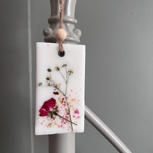 May include: A rectangular white wax melt with a dried red rose and small white flowers. The wax melt is adorned with dried petals and a wooden bead, suspended by a natural twine cord. The background is a soft gray.