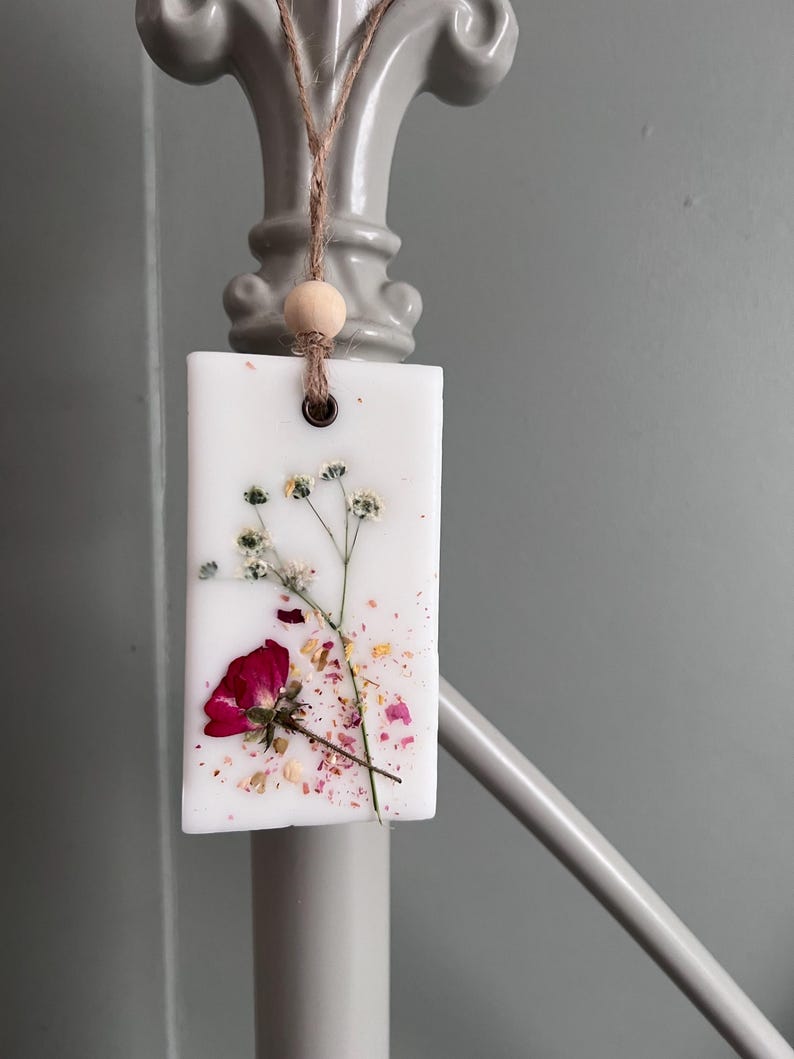 May include: A rectangular white wax melt with a dried red rose and small white flowers. The wax melt is adorned with dried petals and a wooden bead, suspended by a natural twine cord. The background is a soft gray.
