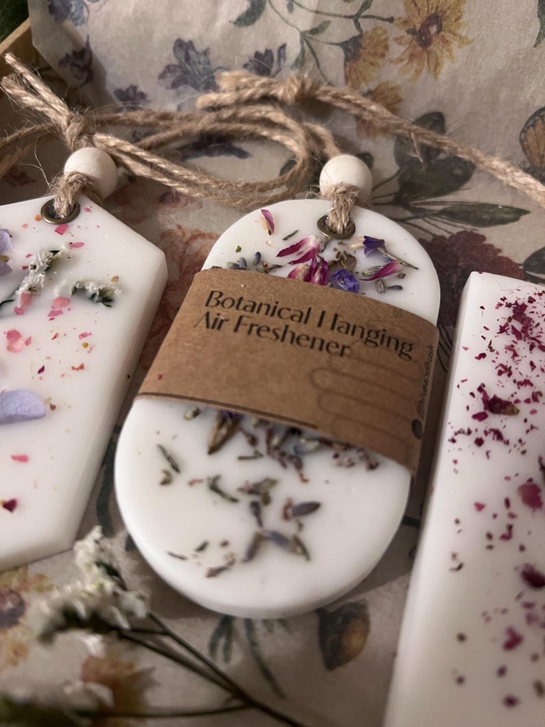 May include: Close-up of three white botanical hanging air fresheners with dried flowers and twine. The center freshener has a brown label that reads "Botanical Hanging Air Freshener". The fresheners are in a floral-patterned box.