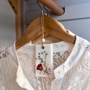 May include: A white lace shirt hangs on a wooden hanger. A rectangular, white wax sachet with dried flowers and gold flakes is suspended from the hanger. The sachet features a red rose and sprigs of delicate greenery.