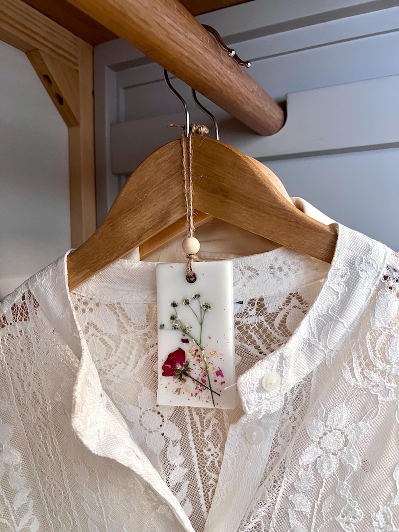 May include: A white lace shirt hangs on a wooden hanger. A rectangular, white wax sachet with dried flowers and gold flakes is suspended from the hanger. The sachet features a red rose and sprigs of delicate greenery.