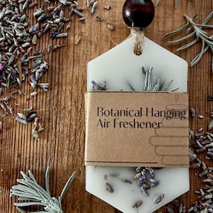 May include: A white, hexagonal botanical hanging air freshener with lavender buds and sprigs. The air freshener is suspended by a jute string and a dark wooden bead. The words "Botanical Hanging Air Freshener" are printed on a brown label.