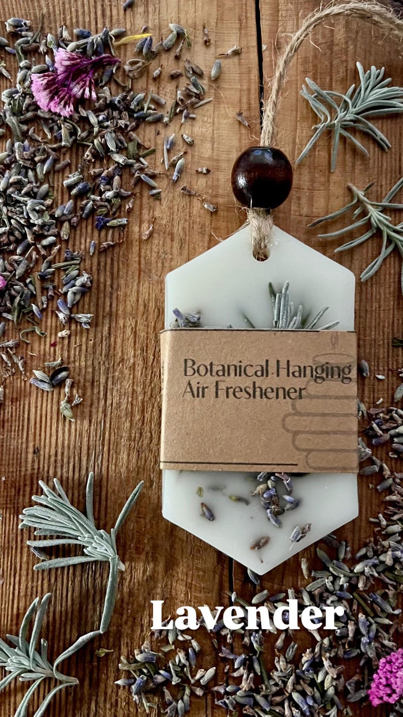 May include: A white, hexagonal botanical hanging air freshener with lavender buds and sprigs. The air freshener is suspended by a jute string and a dark wooden bead. The words "Botanical Hanging Air Freshener" are printed on a brown label.