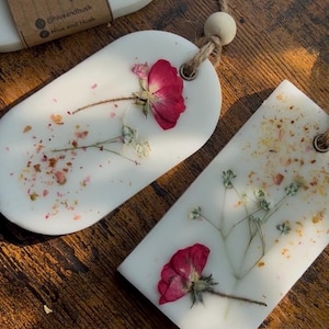 May include: White botanical hanging air fresheners with dried flowers and petals. The air fresheners are oval and rectangular, with a natural twine string and wooden bead for hanging. The image shows the product on a wooden surface.