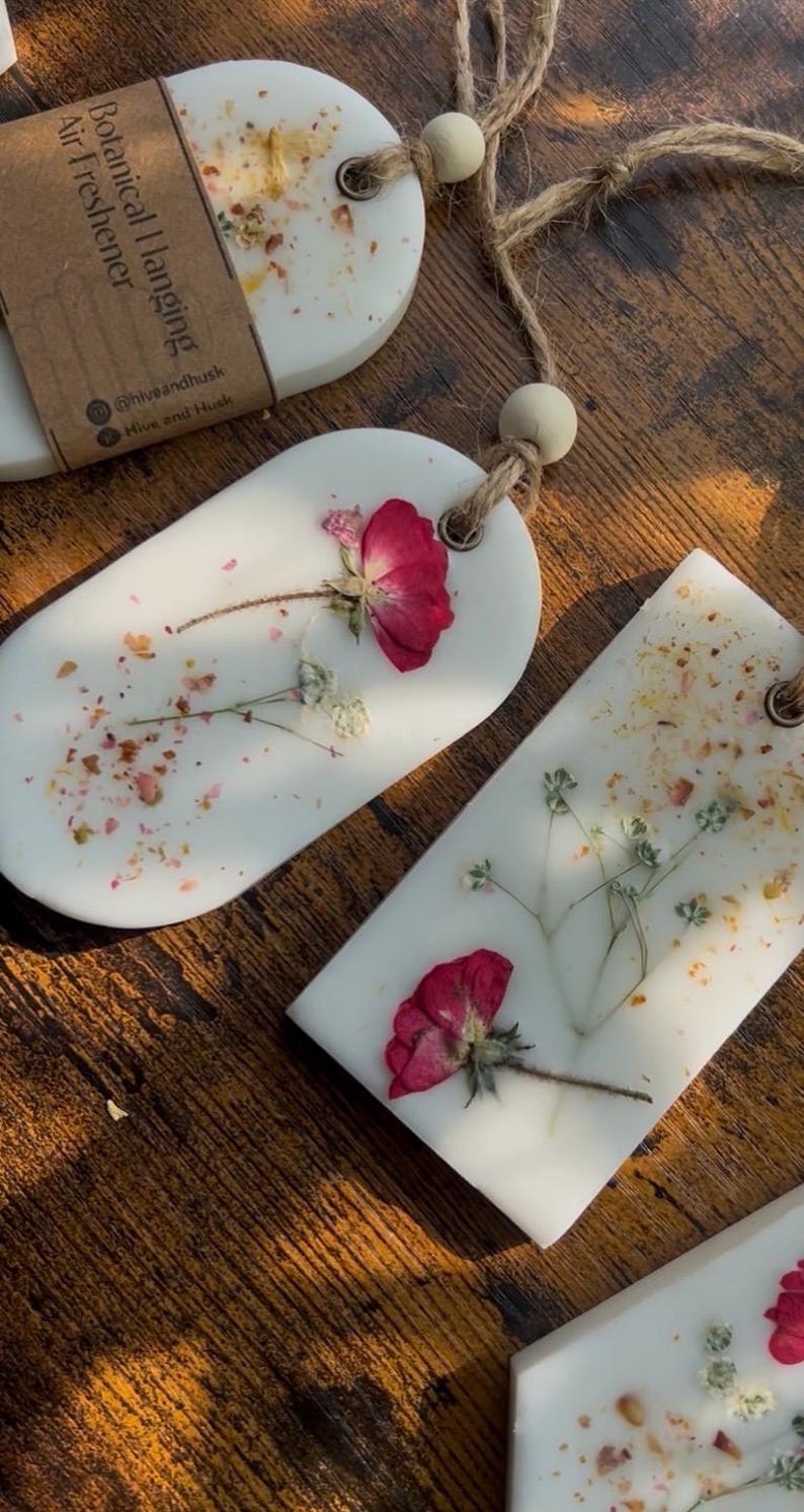 May include: White botanical hanging air fresheners with dried flowers and petals. The air fresheners are oval and rectangular, with a natural twine string and wooden bead for hanging. The image shows the product on a wooden surface.