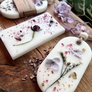 May include: White, oval-shaped wax melts with dried flowers and petals embedded in them. Some melts have a small hole for hanging. The melts are on a wooden surface, with additional dried flowers and greenery.