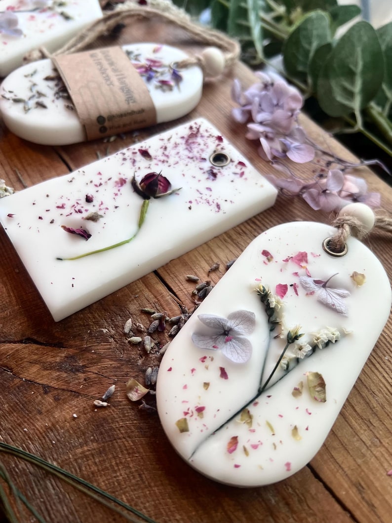 May include: White, oval-shaped wax melts with dried flowers and petals embedded in them. Some melts have a small hole for hanging. The melts are on a wooden surface, with additional dried flowers and greenery.
