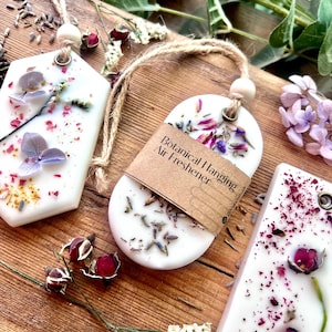 May include: Three white botanical hanging air fresheners with dried flowers and herbs. One is oval-shaped with a brown paper label that reads "Botanical Hanging Air Freshener". The other two are rectangular and hexagonal. All have a jute string for hanging.
