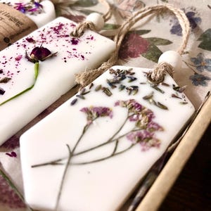 May include: Close-up of several white, geometric-shaped wax melts with dried flowers and herbs embedded on top. Each melt has a jute string for hanging. The background features floral patterned paper.