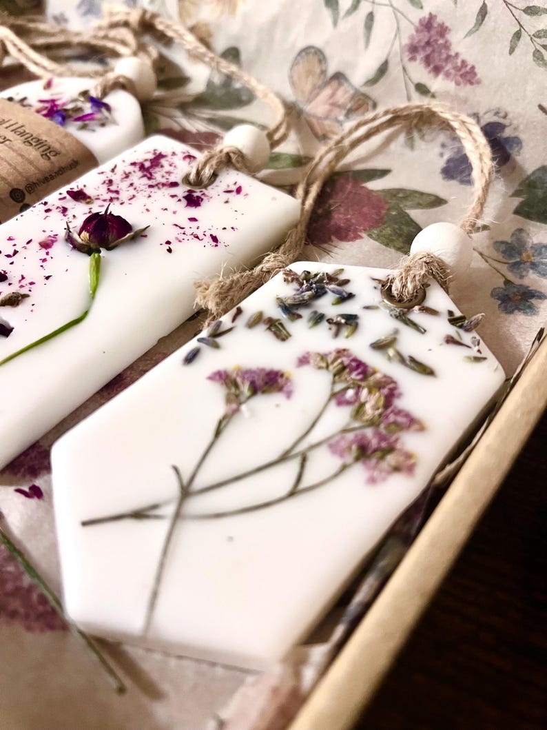 May include: Close-up of several white, geometric-shaped wax melts with dried flowers and herbs embedded on top. Each melt has a jute string for hanging. The background features floral patterned paper.