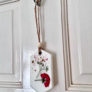 May include: A white, hexagonal-shaped air freshener with a red flower and dried floral accents. The air freshener hangs from a metal hook with a natural twine and wooden bead.