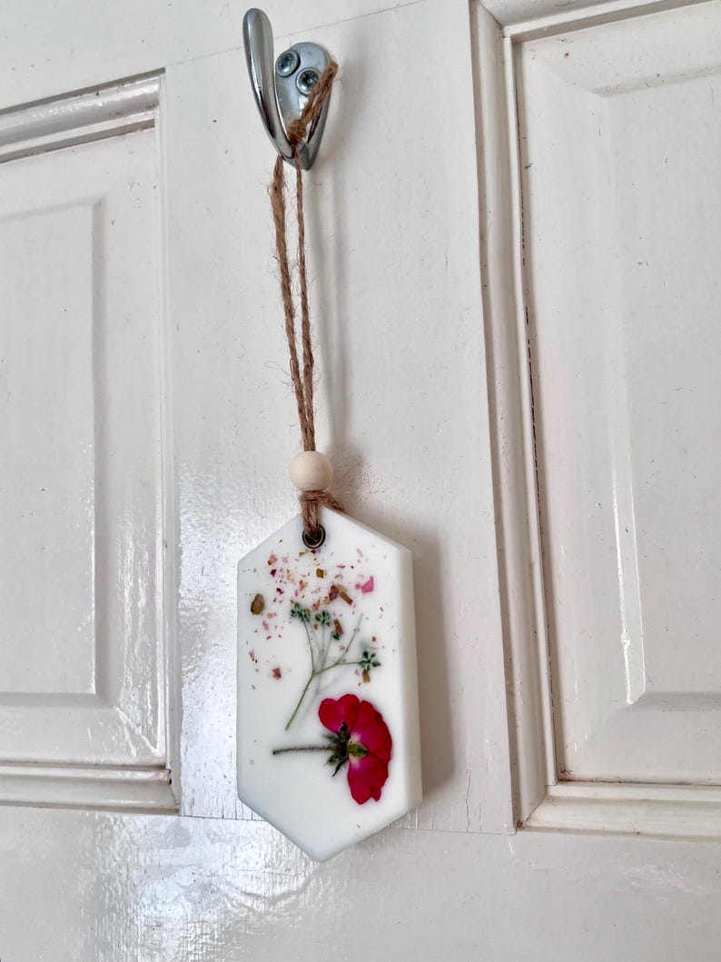 May include: A white, hexagonal-shaped air freshener with a red flower and dried floral accents. The air freshener hangs from a metal hook with a natural twine and wooden bead.